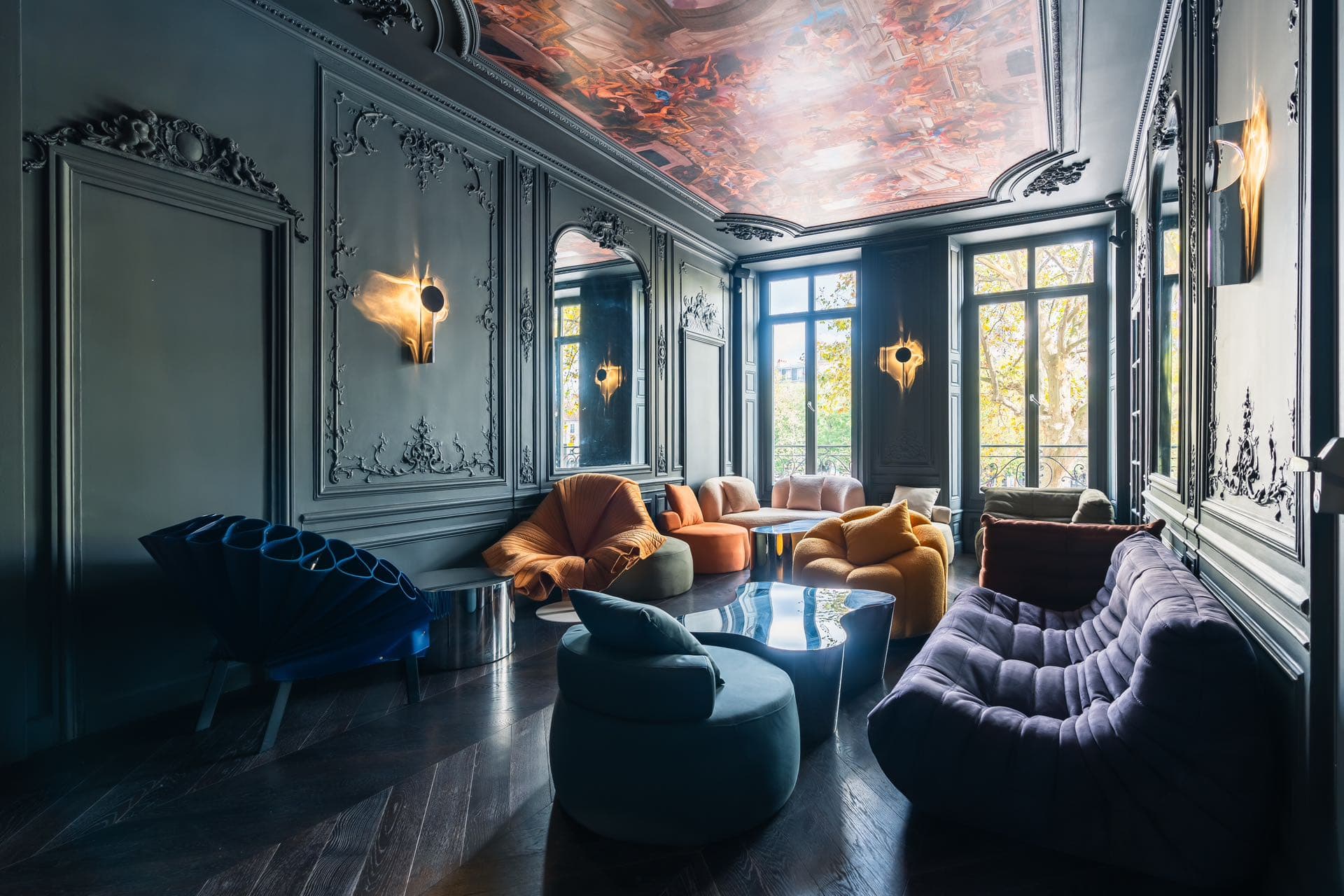 Salon aux boiseries sombres, plafond peint, fauteuils colorés et tables basses métalliques près de grandes fenêtres